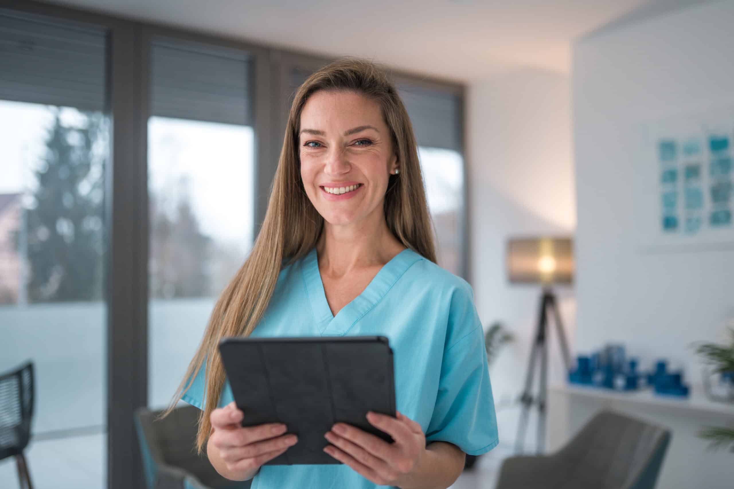 Professional Caucasian female nurse in scrubs holding a digital tablet and looking at the camera with a confident smile inside a well-lit medical facility.