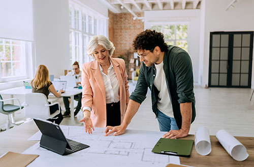 Two colleagues standing together, engaged in conversation while reviewing architectural blueprints. 